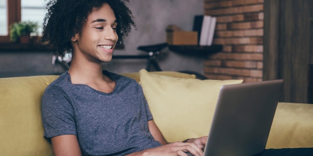 Home work. Cheerful young African man working on laptop and smiling while sitting on the couch at home