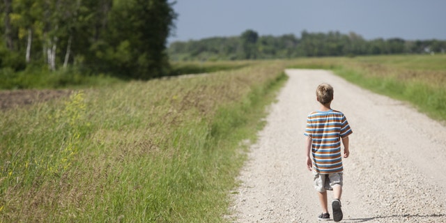 A young boy walks alone down a gravel road in the countryside.