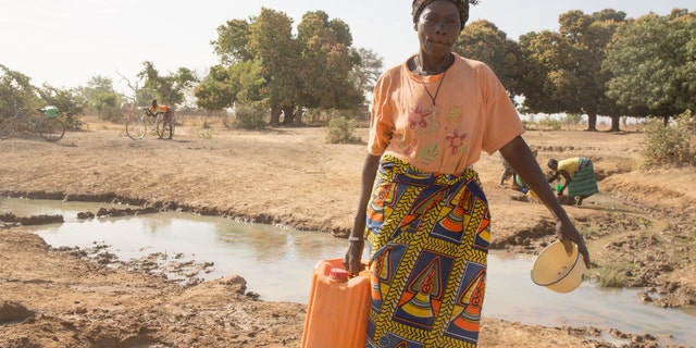 Odette Sompougdo, standing near the dregs of a stream, holding a jerrycan and a dish to collect water from the holes dug around, Vy district in Nassoulou Village, Burkina Faso, January 2016.