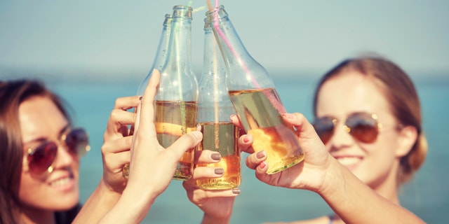 summer vacation, holidays, party, travel and people concept - close up of happy young women with drinks clinking bottles on beach