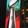 Football fans are dwarfed by a giant projection of the Lombardi trophy on the side of a hotel during Super Bowl LIVE. 