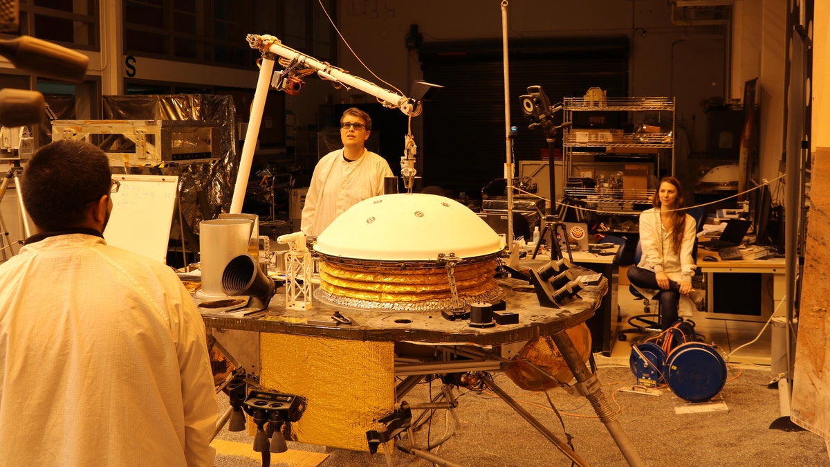 NASA's InSight mission tests an engineering version of the spacecraft's robotic arm in a Mars-like environment at NASA's Jet Propulsion Laboratory. The five-fingered grapple on the end of the robotic arm is lifting up the Wind and Thermal Shield, a protective covering for InSight's seismometer. The test is being conducted under reddish "Mars lighting" to simulate activities on the Red Planet. (Credit: NASA/JPL-Caltech)