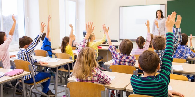 A group of school kids with teacher sitting in classroom and raising hands.
