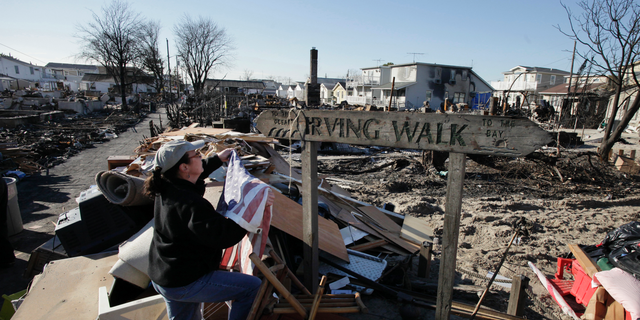 In this Nov. 14, 2012 photo, Louise McCarthy places an American flag on a street sign for Irving Walk in the Breezy Point neighborhood of Queens, N.Y. The sign survived a fire that swept through the seaside community during Superstorm Sandy two weeks earlier. Six years later, most residents have stayed. Homes have been restored, and Sandy led to a new ferry service and a rebuilt boardwalk. (AP Photo/Mark Lennihan)