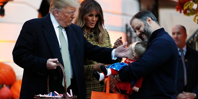 President Donald Trump and first lady Melania Trump greeting a baby in a superhero costume as they hand out candy to children during a Halloween trick-or-treat event at the White House.
