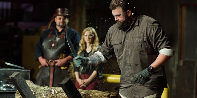 'Mast of Arms' judge Zeke chopping at log with contestants knife. Trent and Ashley in background.