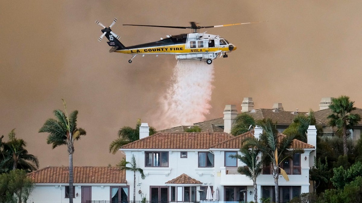 A helicopter drops water on a brush fire behind a home during the Woolsey Fire in Malibu, Calif., Friday, Nov. 9, 2018.