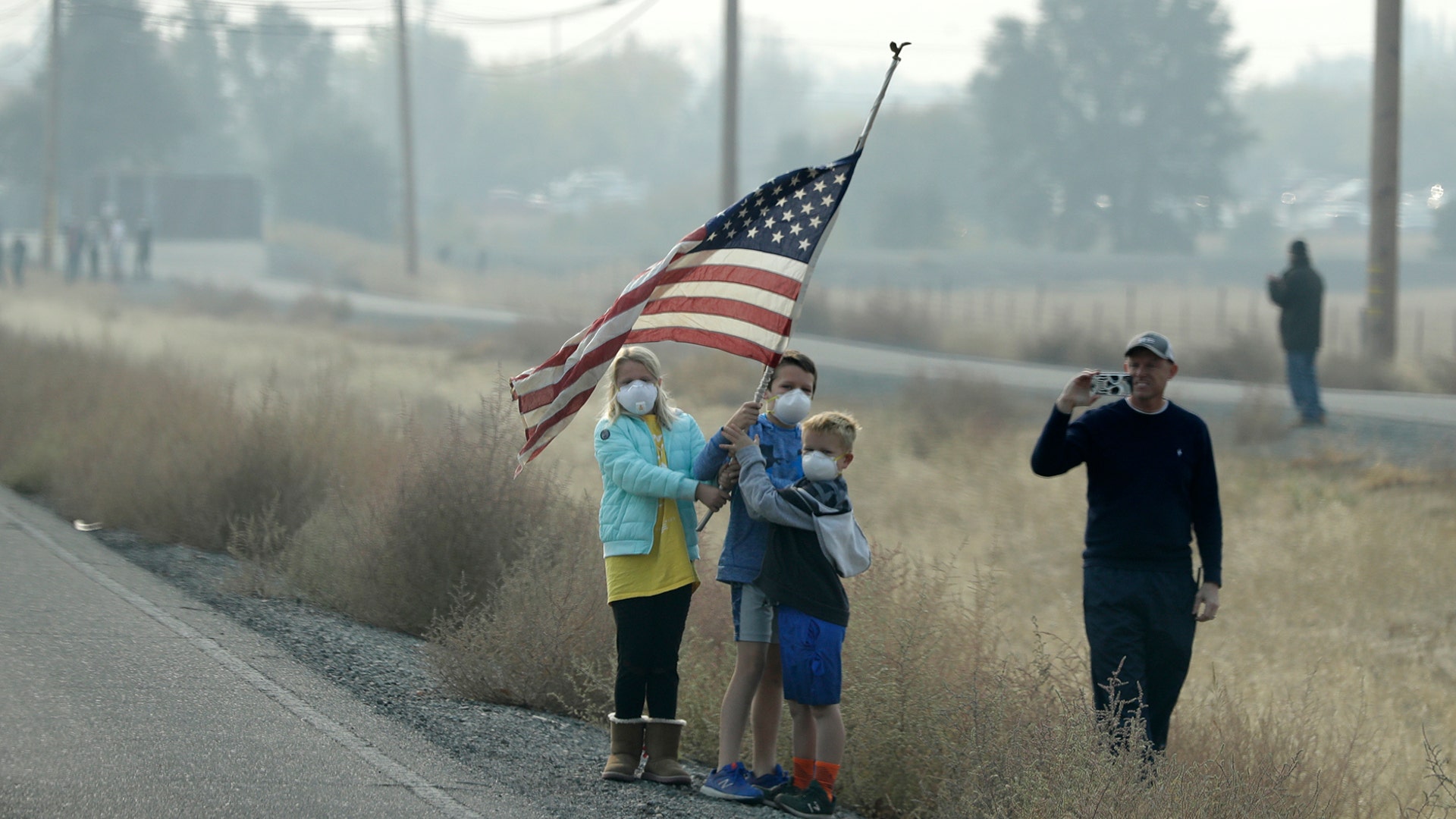 People stand on the side of the road holding an American flag as the motorcade of President Donald Trump drives through areas affected by wildfires in  Chico, California, Nov. 17, 2018. 
