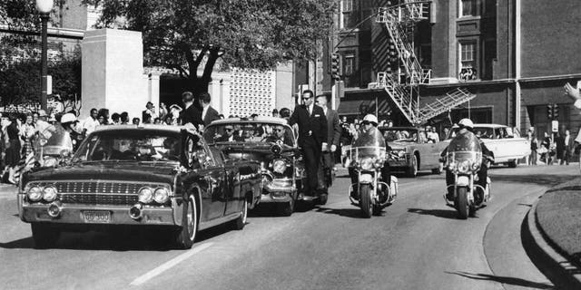 FILE - In this Nov. 22, 1963, file photo, seen through the foreground convertible's windshield, President John F. Kennedy's hand reaches toward his head within seconds of being fatally shot as first lady Jacqueline Kennedy holds his forearm as the motorcade proceeds along Elm Street past the Texas School Book Depository in Dallas.