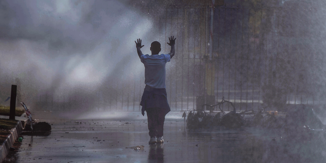 FILE - In this Aug. 1, 2018 file photo an opposition MDC party supporter hold his hands up after being sprayed with a water canon and in clouds of teargas on a street in Harare during clashes with police. Zimbabweans have reacted with stunned disbelief to the testimony from two generals who denied troops killed six people in August when the military was called in to crush protests following the country’s disputed elections. (AP Photo/Mujahid Safodien, File)