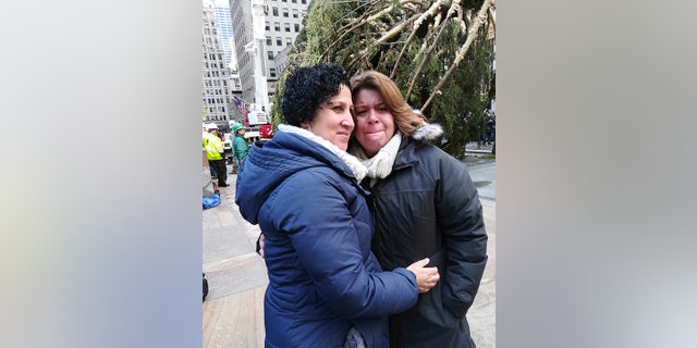Lissette Gutierrez, left, hugs her wife, Shirley Figueroa, with the 72-foot-tall Norway spruce that the couple donated in the background, Saturday, Nov. 10, 2018 in New York. The tree was hoisted into position by crane after making the 60-mile trip from the couple's Wallkill, New York, home and will become the 2018 Rockefeller Center Christmas tree. (AP Photo/Julie Walker)