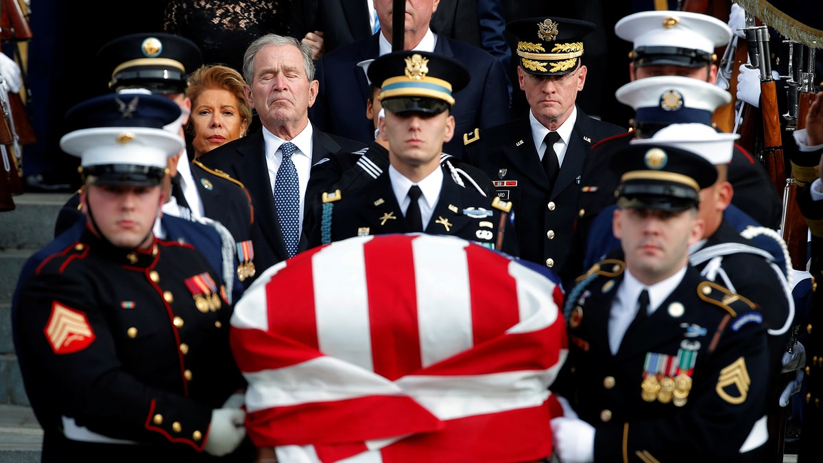 Former President George W. Bush closes his eyes in solemn reflection as military pallbearers carry the flag-draped casket of former President George H. W. Bush during a state funeral ceremony attended by family members, military officials, and dignitaries.