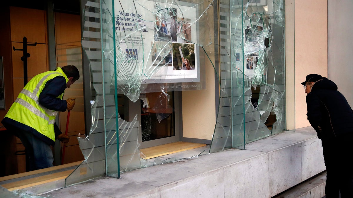 A worker clears debris in a bank as a man watches through smashed windows, in Paris, Sunday, Dec. 9, 2018. (AP Photo/Christophe Ena)