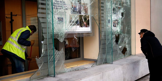 A worker clears debris in a bank as a man watches through smashed windows, in Paris, Sunday, Dec. 9, 2018. (AP Photo/Christophe Ena)