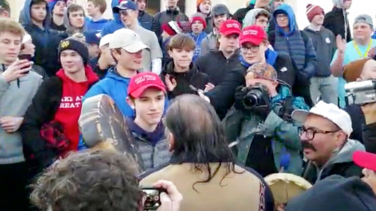 In this Friday, Jan. 18, 2019 image made from video provided by the Survival Media Agency, a teenager wearing a "Make America Great Again" hat, center left, stands in front of a Native American who approached him singing and playing a drum in Washington. (Survival Media Agency via AP)
