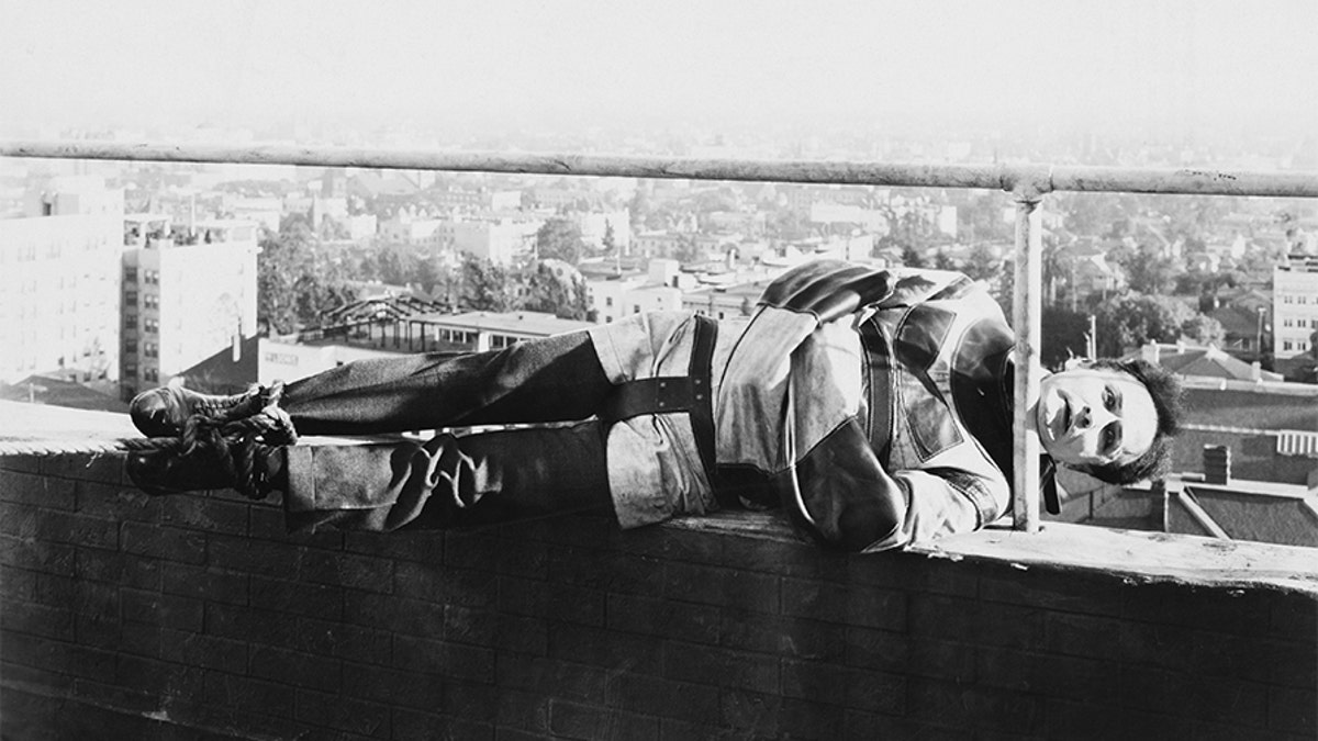 Harry Houdini holds onto a railing by his chin while tied up in a straight jacket.