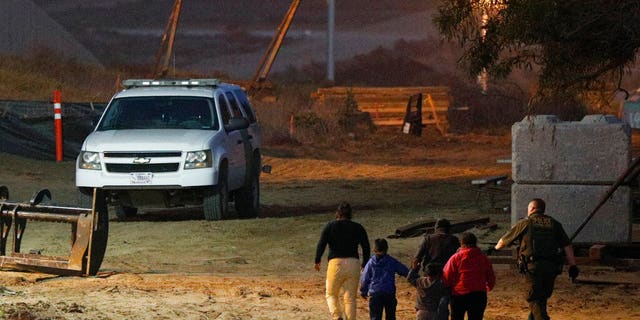 FILE - In this Dec. 3, 2018, file photo, migrants are escorted by a U.S. Border Patrol agent as they are detained after climbing over the border wall from Playas de Tijuana, Mexico, to San Ysidro, Calif. (AP Photo/Rebecca Blackwell, File)