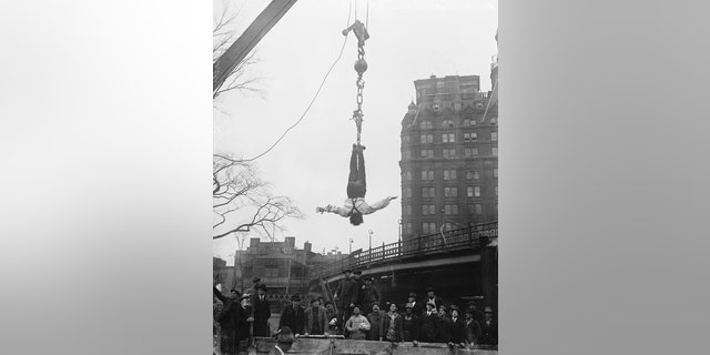 Harry Houdini hangs upside down from a crane after freeing himself from a straitjacket.