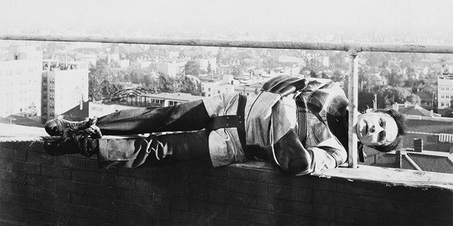 Harry Houdini holds onto a railing by his chin while tied up in a straight jacket.