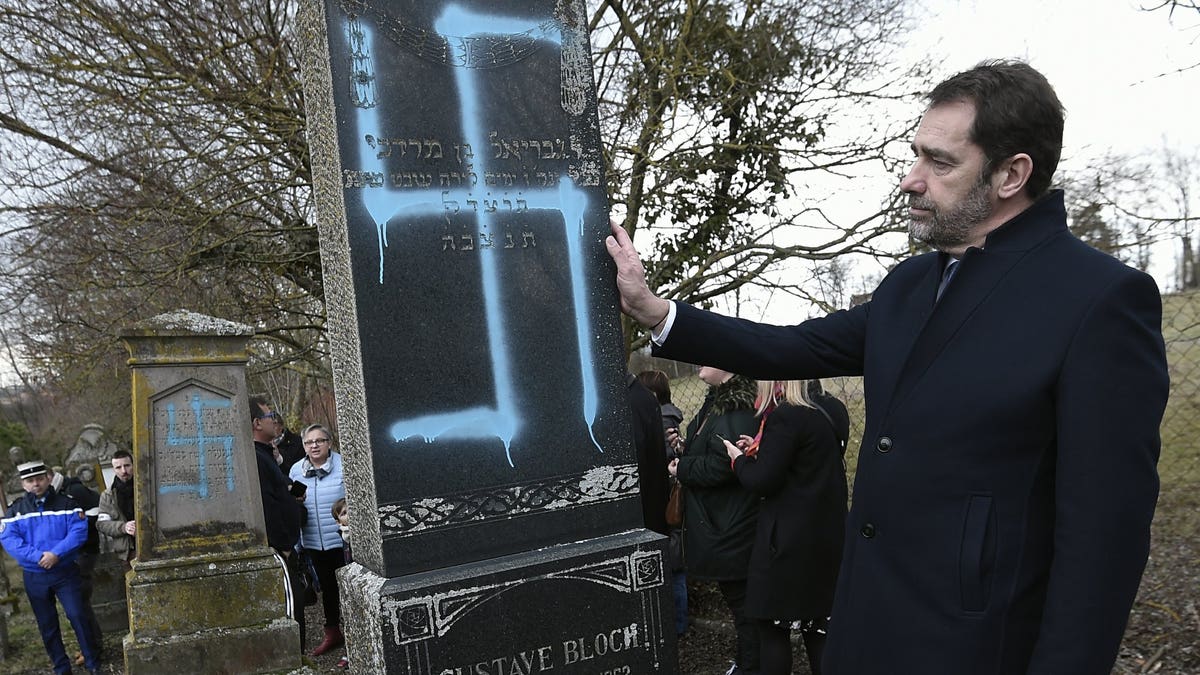 French Interior Minister Christophe Castaner touches a vandalized tombstone at the Jewish cemetery of Quatzenheim, eastern France, Tuesday Feb. 19, 2019. French residents and public officials from across the political spectrum geared up Tuesday for nationwide rallies against anti-Semitism following a series of anti-Semitic acts, including the swastikas painted on about 80 gravestones at the Jewish cemetery overnight.