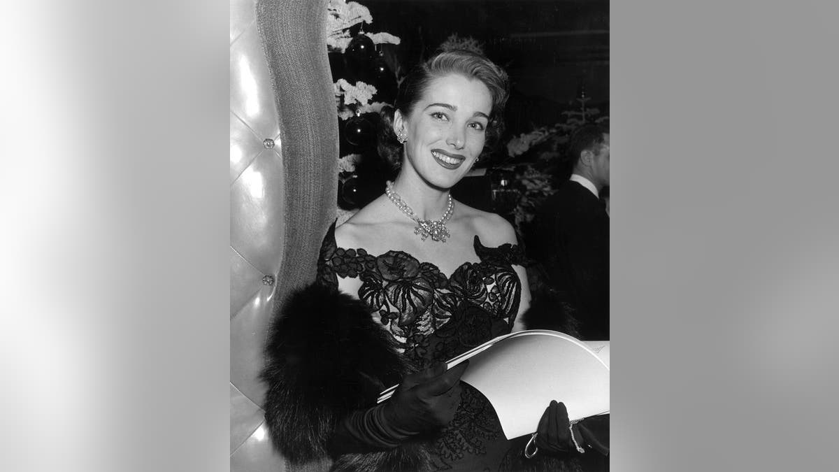 Circa 1950: A portrait of American actor Julie Adams wearing a strapless evening gown, pearls, and long gloves while holding a menu in a restaurant.