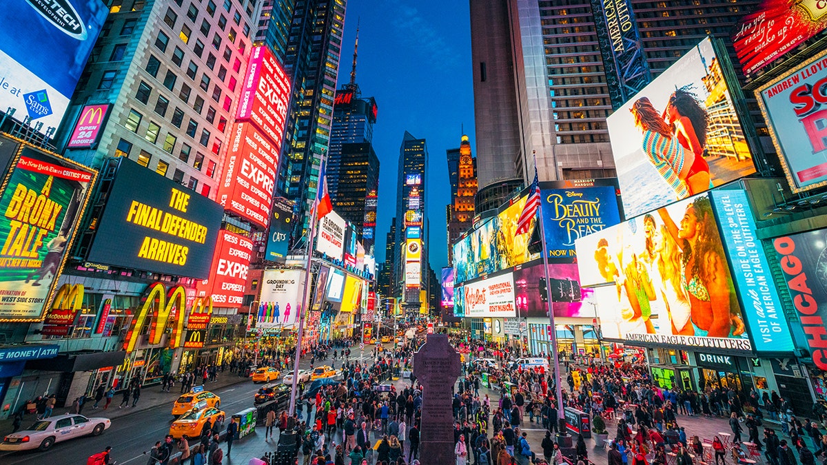 Times Square covered with illuminated billboards during the 'blue hour' period at dusk, with tourists on the streets.