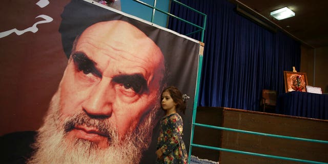 In this Tuesday, Jan. 22, 2019, photo, a girl walks past a poster of Ayatollah Ruhollah Khomeini, at a mosque where he made speeches, in northern Tehran, Iran. The memory of Khomeini, who died in 1989 at the age of 86, looms large over Tehran today. His image is on bank notes and in textbooks in Iran, often as an embodiment of the 1979 Islamic Revolution that swept aside the country’s shah and forever changed the nation.
