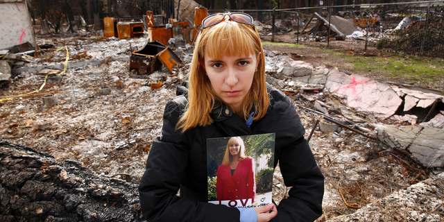 In this Thursday, Feb. 7, 2019 photo, Christina Taft, the daughter of Camp Fire victim Victoria Taft, poses with a photo of her mother, at the burned out ruins of the Paradise, Calif., home where she died last fall. Taft refused to leave. If the threat was real, authorities would order an evacuation, she told her daughter. Victoria Taft’s remains were recovered from the ruins of her living room. (AP Photo/Rich Pedroncelli)