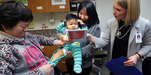 CORRECTS SPELLING OF ABEL, NOT ABLE - As his mother Wenyi Zhang holds him, one-year-old Abel Zhang looks at the book being given him by Dr. Lauren Lawler, right, as his grandmother Ding Hong helps with his clothes moments after the child received the last of three inoculations, including a vaccine for measles, mumps, and rubella (MMR), at the International Community Health Services Wednesday, Feb. 13, 2019, in Seattle. A recent measles outbreak has sickened dozens of people in the Pacific Northwest, most in Washington state and, of those, most are concentrated in Clark County, just north of Portland, Oregon. Washington Gov. Jay Inslee declared a state of emergency over the outbreak last month. (AP Photo/Elaine Thompson)