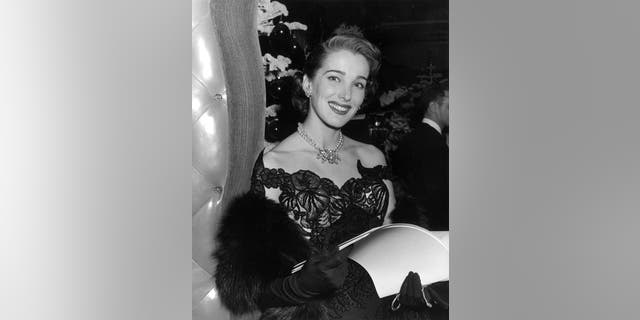 Circa 1950: A portrait of American actor Julie Adams wearing a strapless evening gown, pearls, and long gloves while holding a menu in a restaurant.
