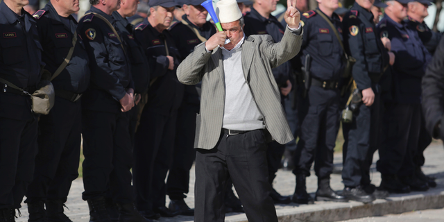 A protester flashes the victory sign in front of Albanian policemen guarding the parliament building as opposition supporters gather to take part in an anti-government rally , in Tirana , Albania, Tuesday, March 5, 2019. The opposition demanded the government step down, claiming it's corrupt and has links to organized crime. (AP Photo/ Hektor Pustina)