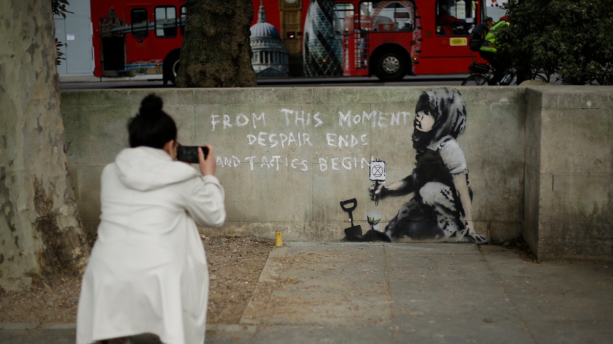 A woman takes a picture of a new piece of street art that people noticed for the first time last night and is believed to be by street artist Banksy on a wall where Extinction Rebellion climate protesters had set up a camp in Marble Arch, London, Friday, April 26, 2019. 