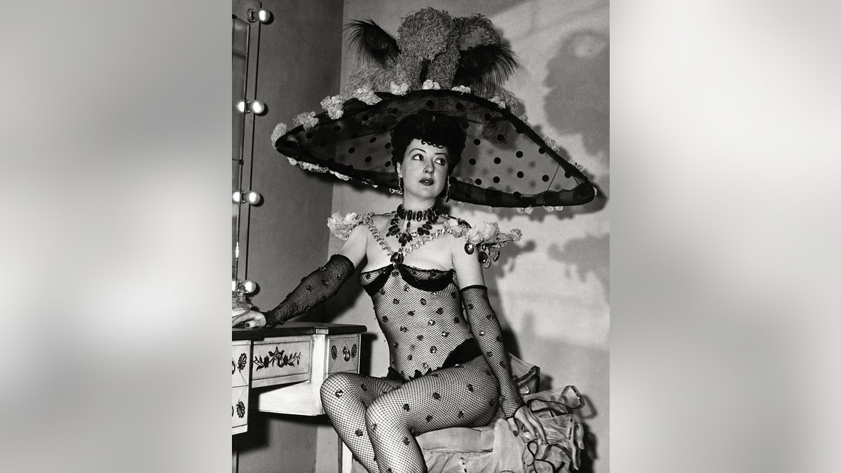 Gypsy Rose Lee wearing a one-piece fishnet unitard with large sequins, matching gloves and a large feather and netting hat.