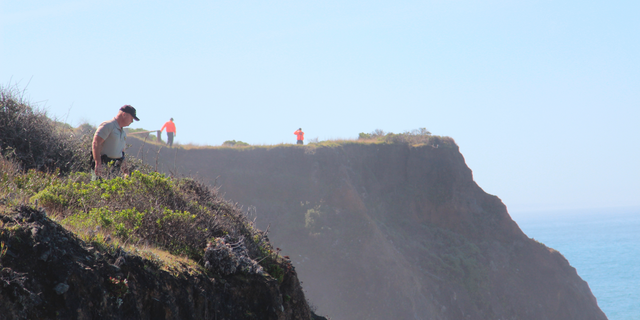 FILE - In this March 29, 2018 file photo, Deputy Bill Holcomb looks down the cliff near the crash site near Mendocino, Calif., as search and rescue volunteers scour the area behind him on and resume looking for children, still missing after their parent's SUV plunged into the ocean Monday. A California Highway Patrol investigator testified Thursday, April 4, 2019 that Sarah Hart searched whether death by drowning was relatively painless hours before her wife drove an SUV off a cliff, killing them and six adopted children in waters below. A special coroner's jury in Mendocino County is trying to determine whether the March 2018 deaths were murder-suicide or accidental. (Kale Williams/The Oregonian via AP)