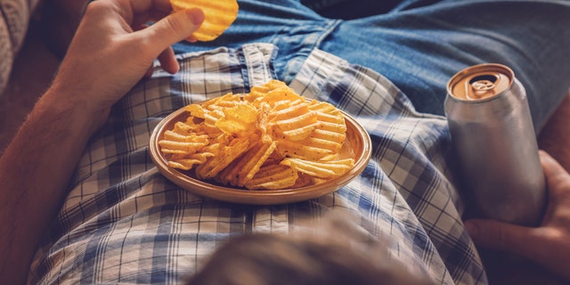 A man lies on a sofa with a beer in his hand — snacking on potato chips balanced on his stomach.