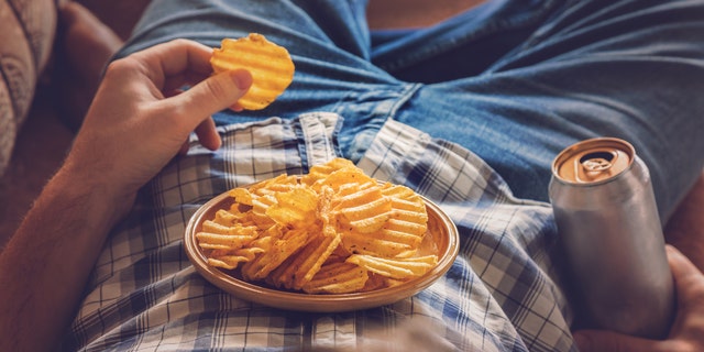 A man lies on the couch with a beer in his hand — while snacking on potato chips balanced on his stomach.