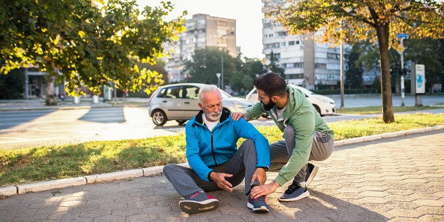 Son helping his father to stand up after his fall while jogging outdoors in city street.