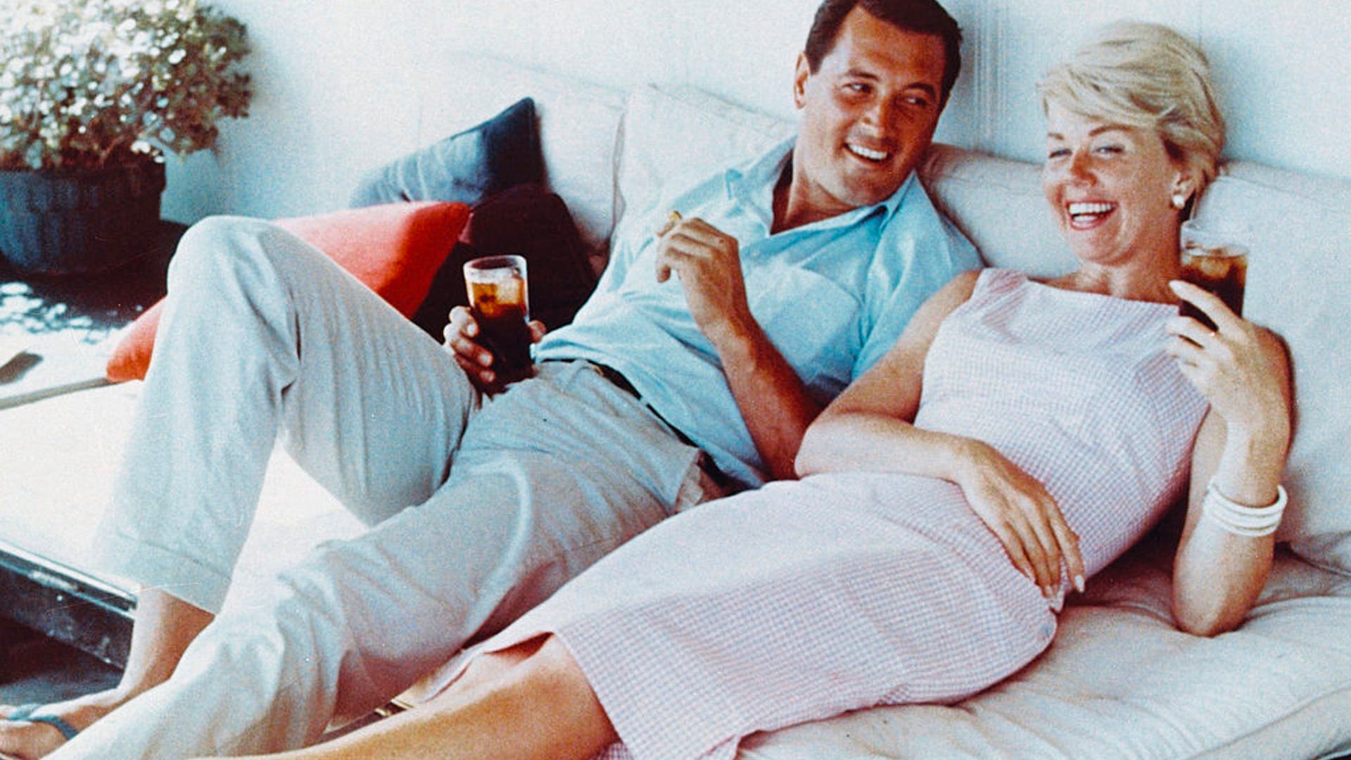 Rock Hudson (1925-1985), US actor, wearing white trousers and a light blue short-sleeved shirt, and Doris Day, US singer and actress, in a pink gingham dress, both reclining on a sofa, laughing and holding drinks, circa 1960. (Photo by Silver Screen Collection/Getty Images)