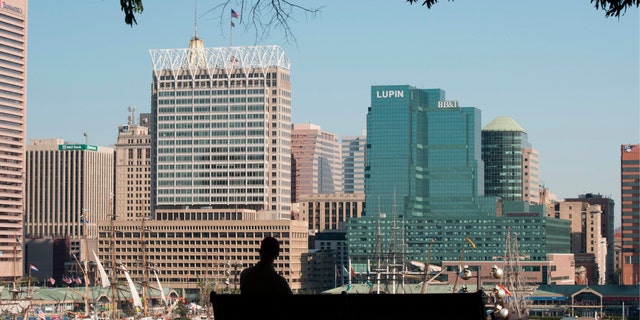 A man sits on a park bench overlooking Baltimore harbor and skyline, Baltimore, Maryland. 