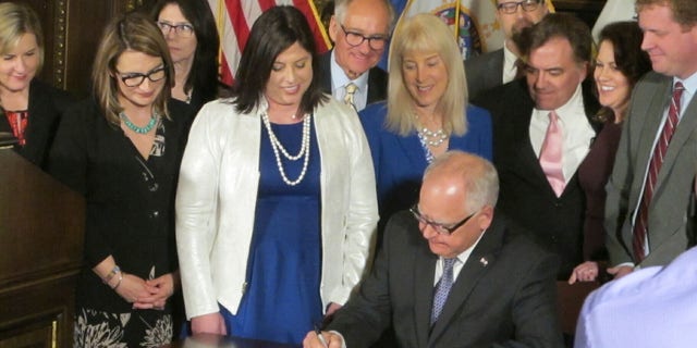 Jenny Teeson, center in white, of Andover, Minnesota, looks on as Minnesota Gov. Tim Walz signs a bill at the Capitol in St. Paul, on Thursday, May 2, 2019, repealing a Minnesota law that prevented prosecutors from filing sexual assault charges against people accused of raping their spouse. Teeson, testified before legislative committees earlier this year about how her now ex-husband drugged her and made a video of himself raping her while she was unconscious. Prosecutors dropped rape charges because of the old law, and he served just 30 days in jail for invasion of privacy. (AP Photo/Steve Karnowski)