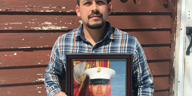 In this April 13, 2019, photo provided by the ACLU of Minnesota, Mark Esqueda poses for a portrait with a photo of him as a U.S.Marine, in Heron Lake, Minn. Esqueda was born in the United States but has been twice denied his request for a passport. He sued the State Department and is asking a federal judge to declare that he is a U.S. citizen. (AP Photo/Lynette Kalsnes, ACLU of Minnesota via AP)