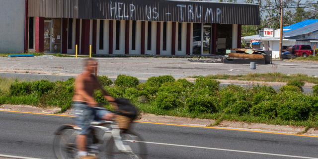 In this May 3, 2019 photo, a cyclist rides by a building damaged by Hurricane Michael in Parker, Fla. Residents in these parts of the Florida Panhandle that were devastated by Hurricane Michael six months ago hope President Donald Trump gets a glimpse of the continuing suffering in the region when he arrives for a campaign rally this week. (AP Photo/Mike Fender)