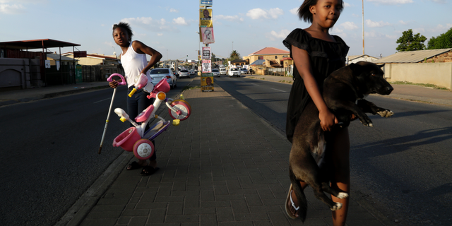 A young girl carries her dog as she crosses the main road, with headline from a newspaper and election posters on the streets of Soweto, South Africa, Friday, May 10, 2019. The ruling African National Congress is coasting to a comfortable lead in South Africa's presidential and parliamentary elections with 80% of the vote counted, but the ongoing tally shows the party getting less support than in the previous poll five years ago. (AP Photo/Themba Hadebe)