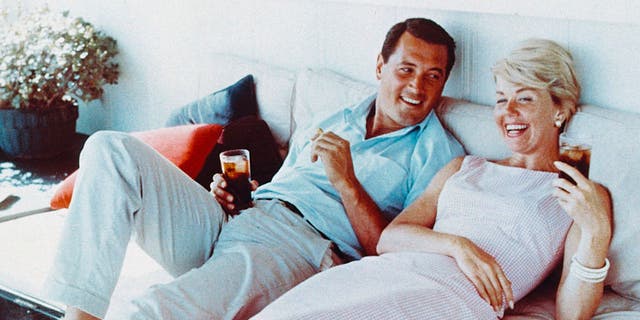 Rock Hudson (1925-1985), US actor, wearing white trousers and a light blue short-sleeved shirt, and Doris Day, US singer and actress, in a pink gingham dress, both reclining on a sofa, laughing and holding drinks, circa 1960. (Photo by Silver Screen Collection/Getty Images)