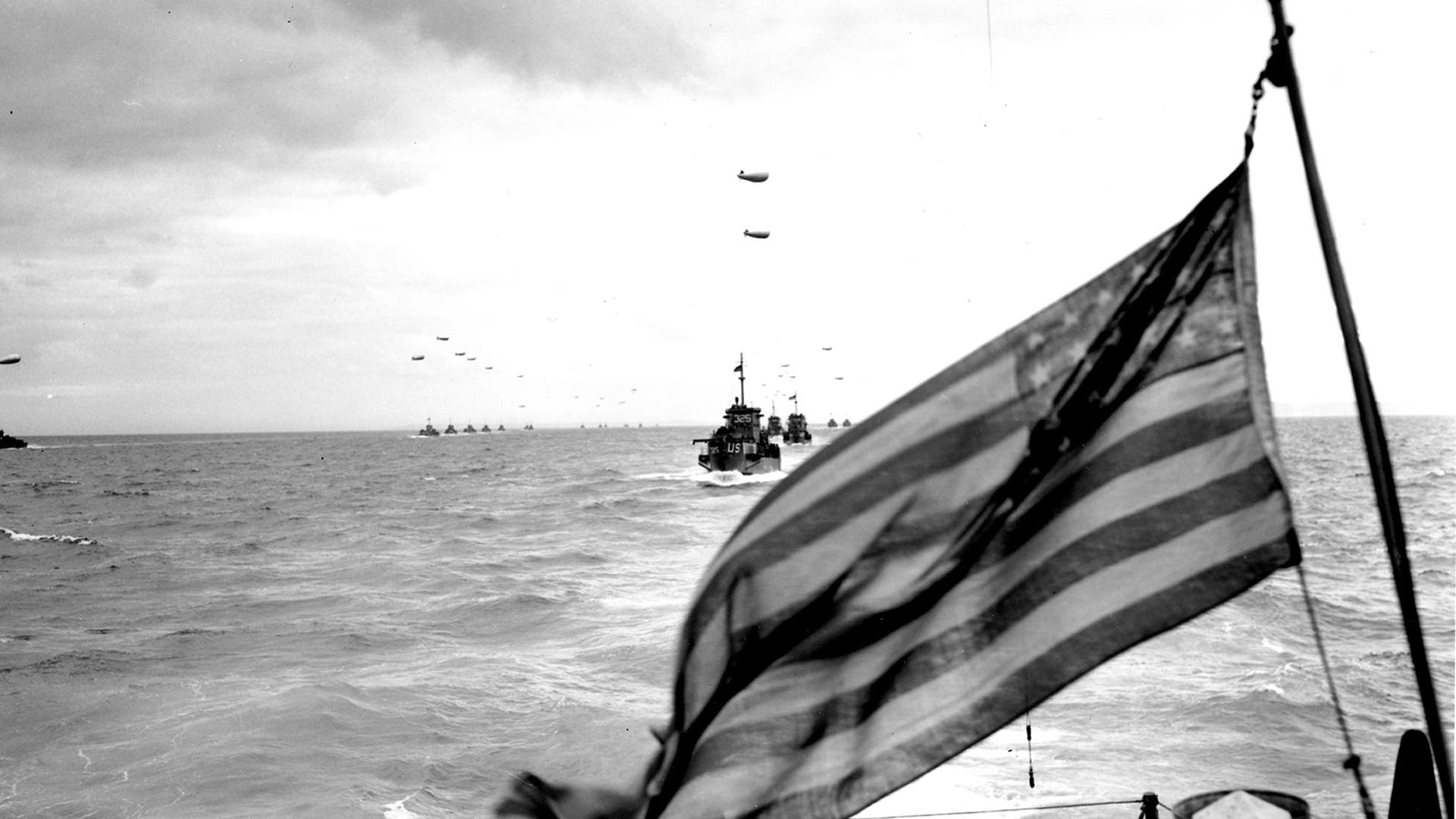 American flag waves through air as war ships sail to Normandy.
