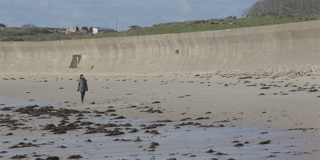 Professor Caroline Sturdy Colls walks along the beach in front of a concrete wall, Alderney.