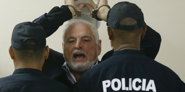 FILE - In this Nov. 19, 2018 file photo, Panama's former President President Ricardo Martinelli answers journalists questions as he raises his handcuffed hands, holding a book, while escorted by police to a hearing at the Supreme Court in Panama City. A Panamanian court has ordered on Wednesday, June 12, 2019, that former President Martinelli to be transferred from jail to house arrest. (AP Photo/Arnulfo Franco, File)