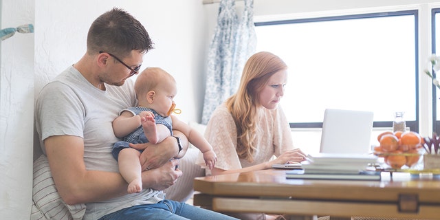 Dad holds her daughter at the kitchen table while mom checks her emails on her laptop next to him with no baby.