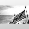 American flag waves through air as war ships sail to Normandy.