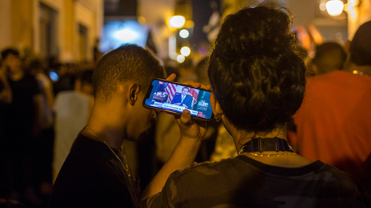 Locals listen on a smartphone to the pre-recorded message by Puerto Rico Gov. Ricardo Rossello announcing that he is resigning Aug. 2 after weeks of protests over leaked obscene, misogynistic online chats, in San Juan, Puerto Rico, Wednesday, July 24, 2019. (Associated Press)